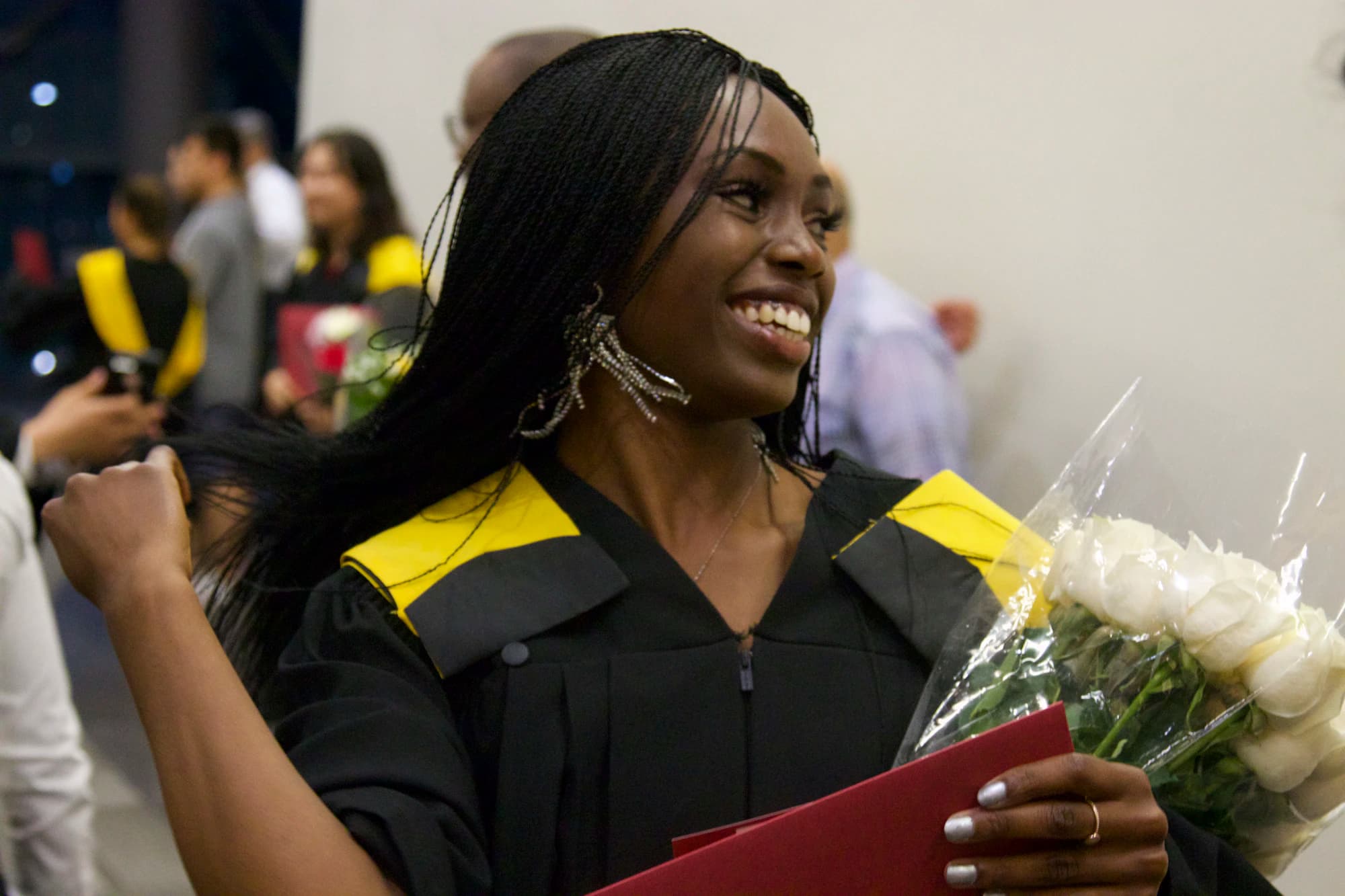 A smiling graduate in a black gown with a yellow stole celebrates, holding a red diploma folder and a bouquet of white roses.