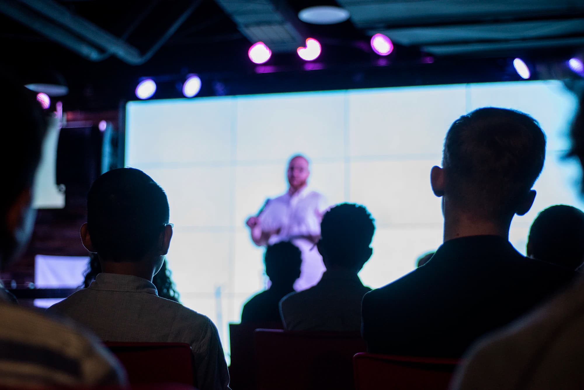 Audience members in silhouette watch a speaker in front of a large glowing screen under purple stage lights.
