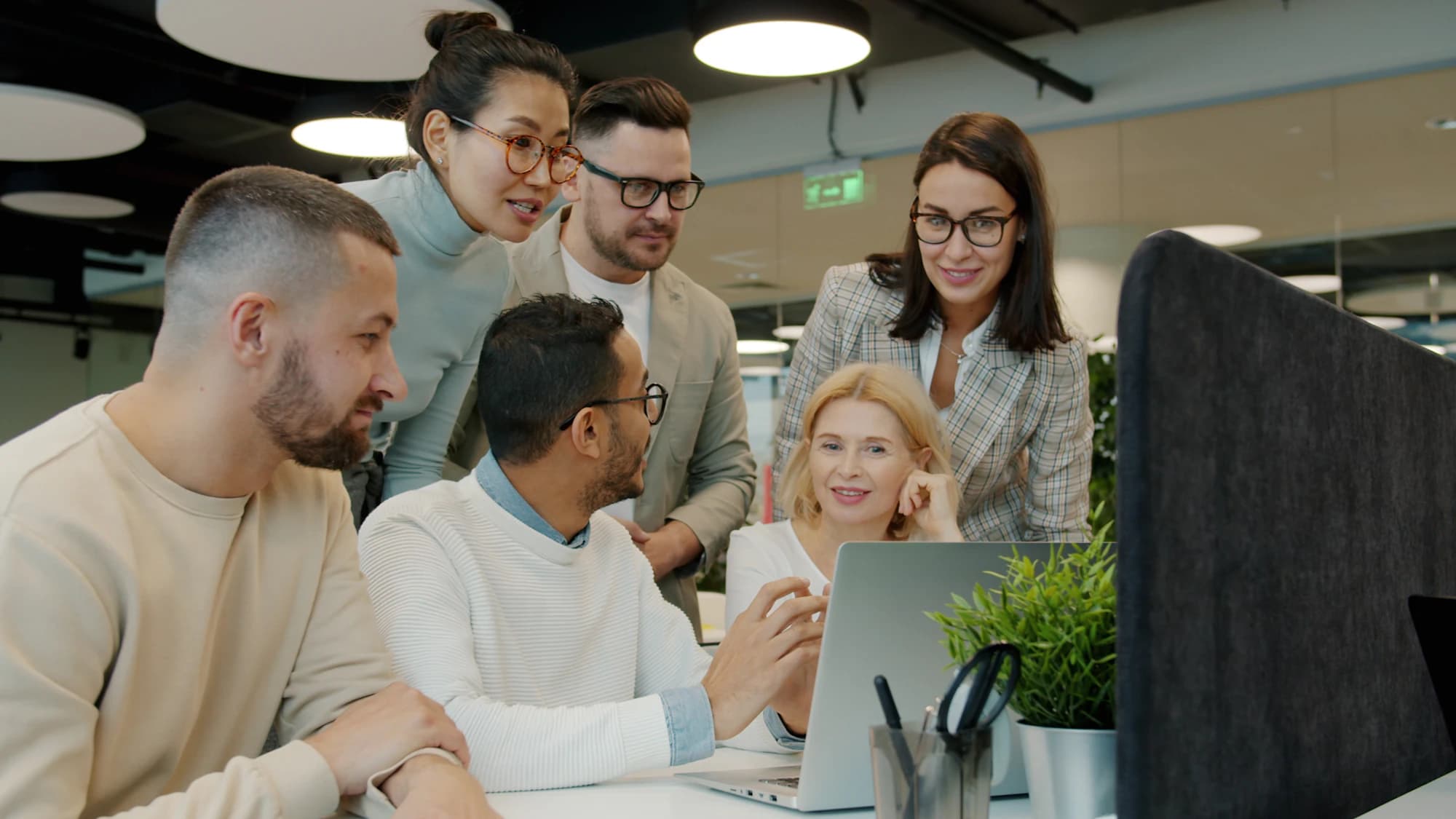 Six colleagues of different ages and backgrounds gather around a laptop at a desk in an open-plan office, discussing what is on screen.